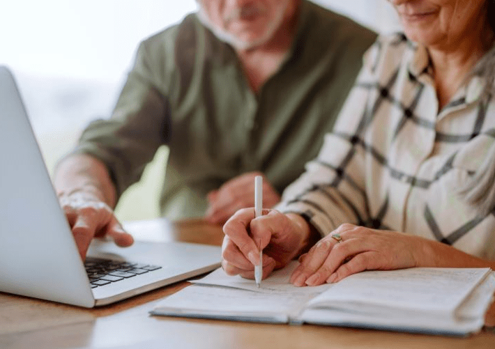A woman writes in a notebook while a man assists her on a laptop. The scene depicts collaboration and learning, likely for a project or study.