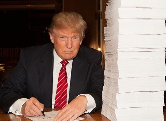 Donald Trump signs documents at a table, with a tall stack of paperwork beside him, illustrating his business dealings or agreements.