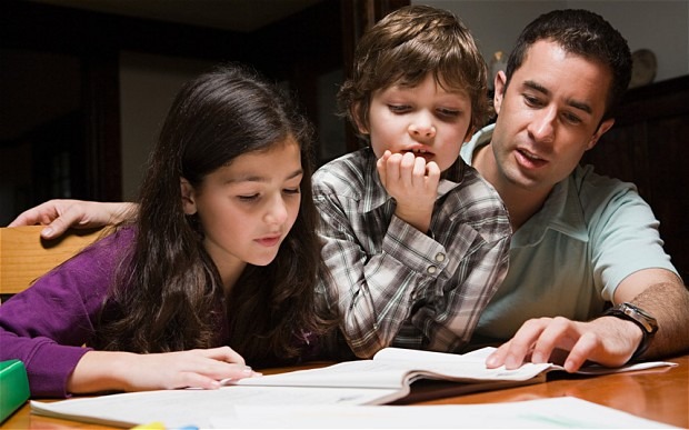 A father helps his two children with their homework, focusing on a book at a table. The scene highlights family learning and support.
