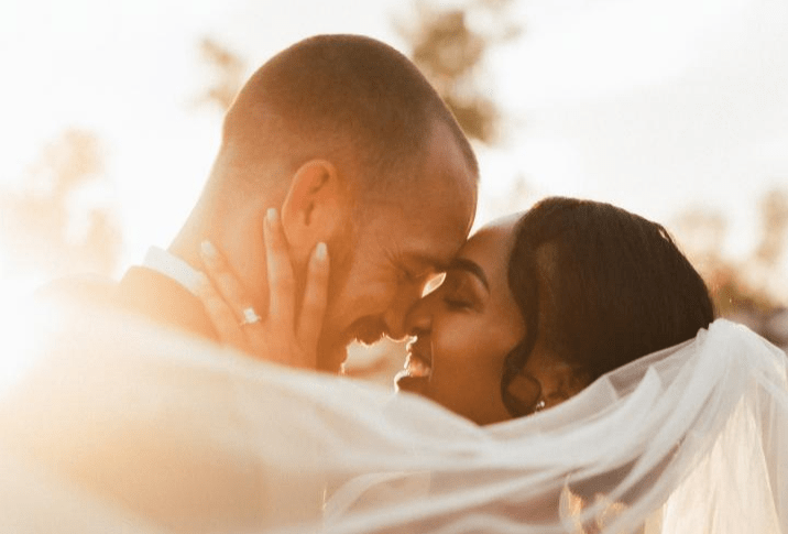 A couple shares a tender kiss, their foreheads touching, surrounded by soft sunlight and flowing veil, capturing a moment of love and intimacy.