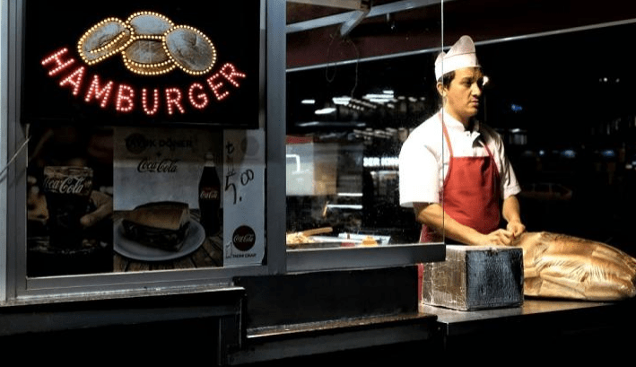 Late-night burger stand featuring a focused cook in a white uniform and red apron, with a glowing "HAMBURGER" sign and displayed menu items.