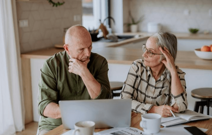 Two older adults engaged in a serious discussion at a kitchen table, with a laptop, coffee cups, and a notepad visible, suggesting planning or problem-solving.