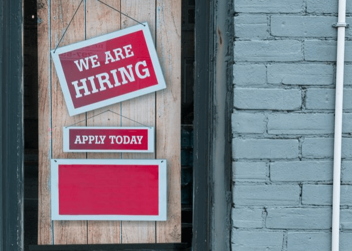 Brightly colored hiring signs read "We Are Hiring" and "Apply Today," inviting job seekers. The signs are displayed on a door against a rustic backdrop.