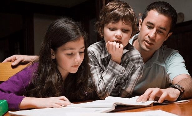 A father helps his two children with reading homework at a table, fostering a supportive learning environment.
