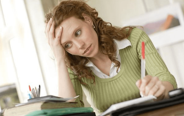 A young woman with curly hair sits at a desk, looking stressed and thoughtful while holding a pen, surrounded by notebooks and office supplies.