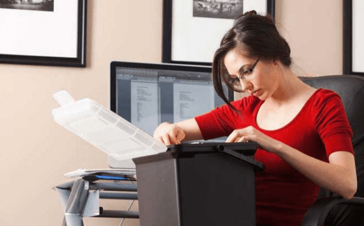 Woman in a red top organizes documents in a black filing box at a tidy home office, with a computer and framed photos in the background.