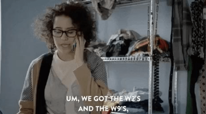 A woman with curly hair and glasses speaks on the phone in a cluttered room, discussing W2 and W9 tax forms.