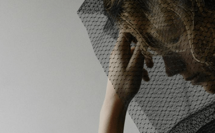 A woman partially obscured by a sheer black veil, gently touching her hair, evokes a sense of intimacy and contemplation against a neutral background.