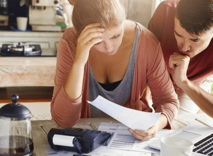 A worried woman examines paperwork at a table, while a man leans in closely, both appearing stressed over financial issues.