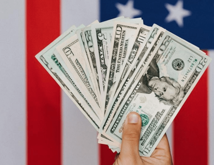 A hand holds several U.S. banknotes in front of an American flag, symbolizing finance and patriotism.