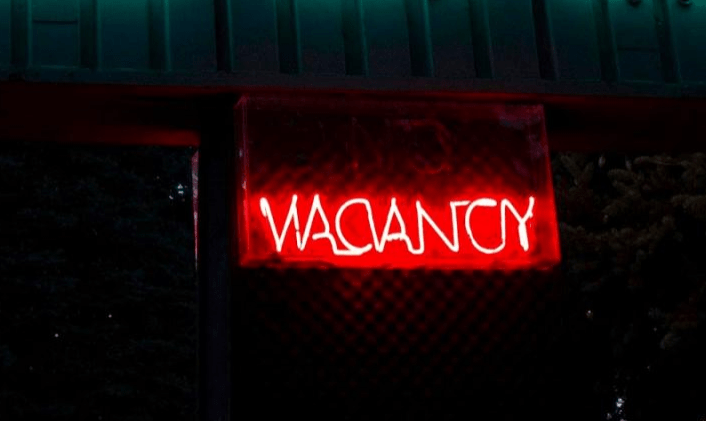 Bright red neon "VACANCY" sign glowing against a dark background, indicating available lodging. Relevant for hotel or travel content.
