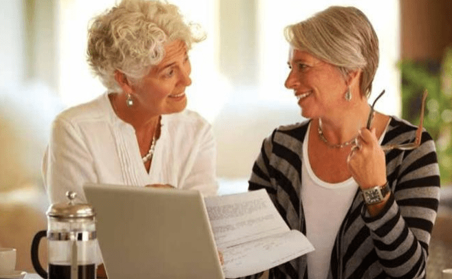 Two smiling older women engage in a lively discussion while reviewing documents next to a laptop, sharing ideas in a warm, inviting setting.