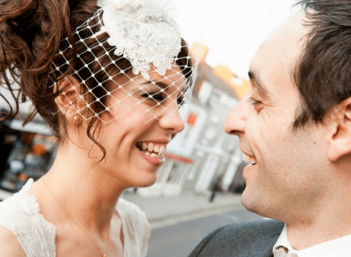 Bride and groom share a joyful gaze, smiling at each other, with the bride wearing a lace veil. A charming street scene sets a romantic backdrop.
