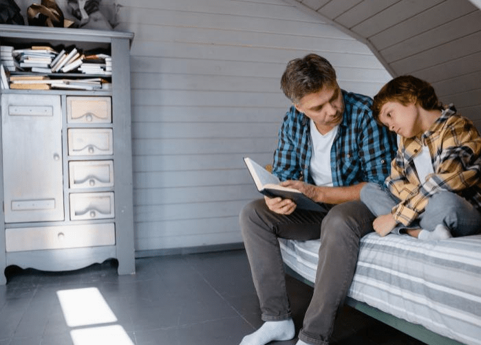 A man reads to a child on a bed in a cozy, well-lit room. Books are stacked nearby, highlighting a nurturing learning environment.