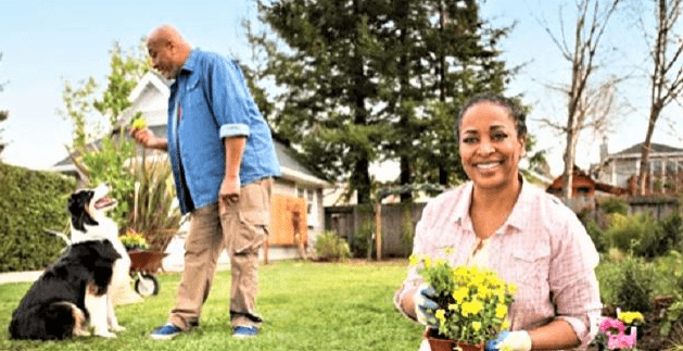 A woman smiles while planting yellow flowers in her garden, as a man stands nearby with a dog, showcasing a vibrant outdoor scene.