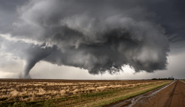 A tornado descends from dark storm clouds over a flat landscape, with a dirt road visible in the foreground, illustrating severe weather conditions.