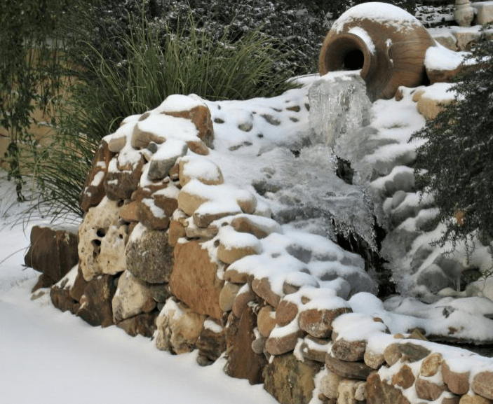 A stone water feature partially covered in snow, with water flowing from a clay pot into a snowy landscape, surrounded by greenery.