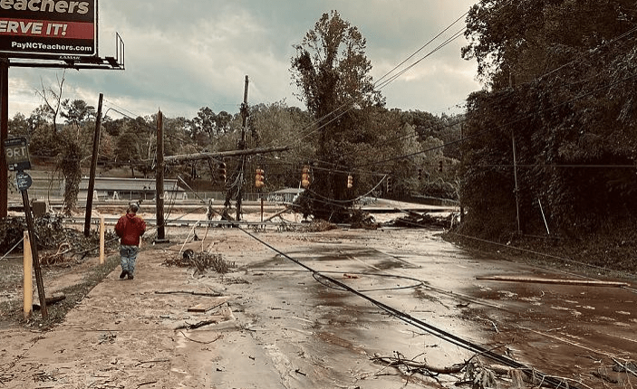 Flooded street with downed power lines and uprooted trees. A lone person walks through debris, highlighting the scene's devastation.