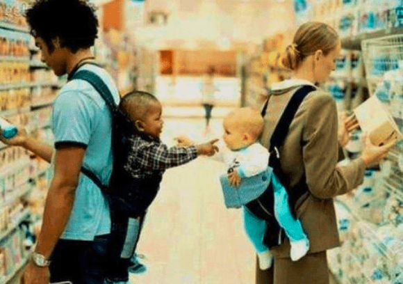 A father and mother shop in a grocery store, carrying a toddler and an infant in baby carriers. The scene highlights parenting and family dynamics.
