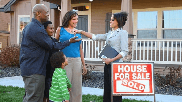 A real estate agent hands keys to a smiling couple outside their newly purchased home, with a "Sold" sign prominently displayed.