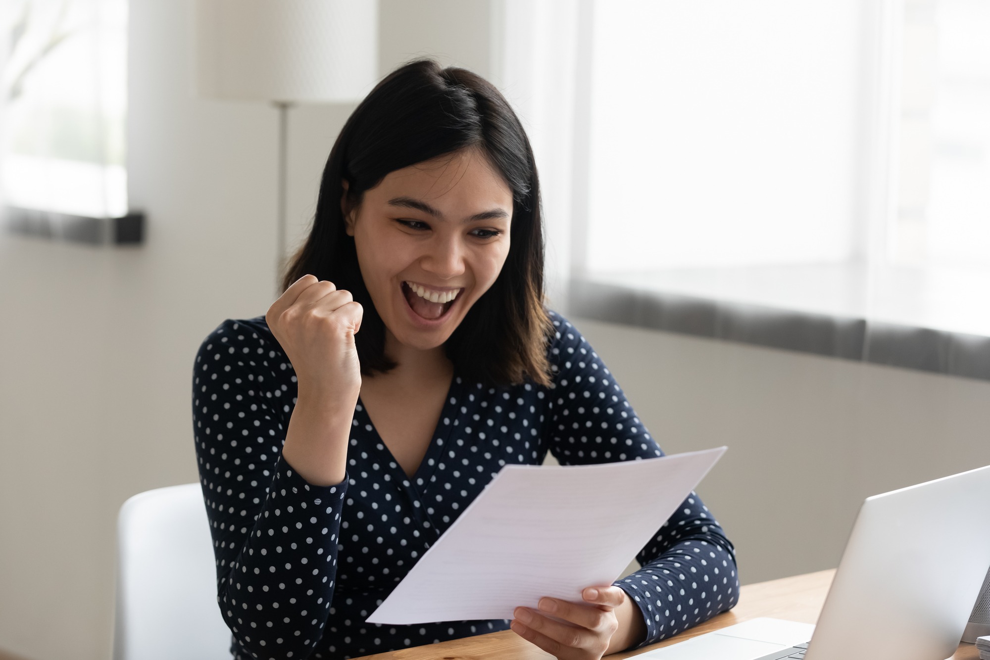 A woman with dark hair, wearing a polka dot shirt, celebrates while holding a document, expressing joy and excitement in a bright, modern workspace.