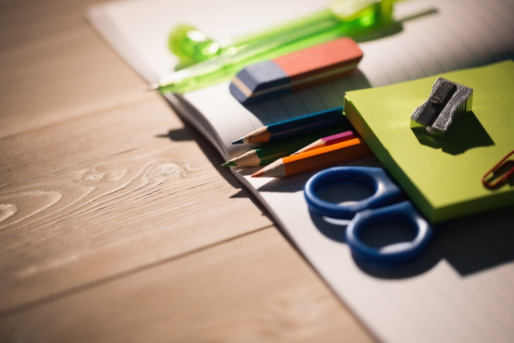 Colorful school supplies, including pencils, scissors, an eraser, and a sharpener, arranged on a wooden desk with a notebook.